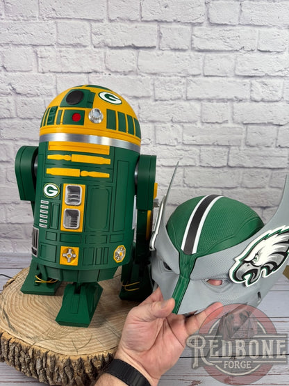 Green and yellow robot figure on a wooden stand with a hand holding a helmet against a brick wall background.